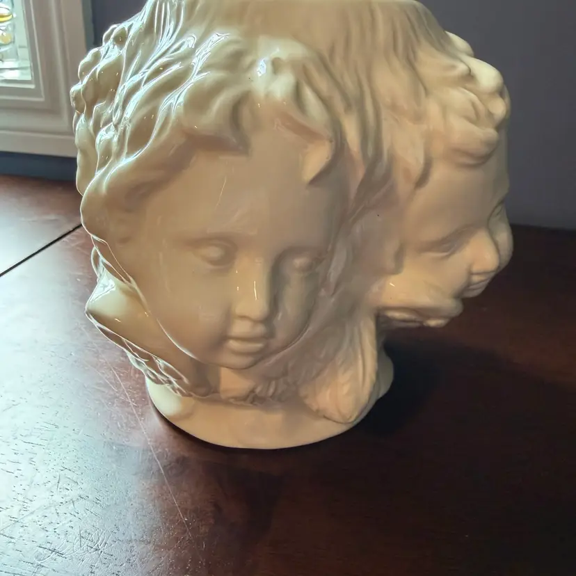 A vintage cream ceramic vase adorned with multiple sculpted cherubic faces. 7” Tall