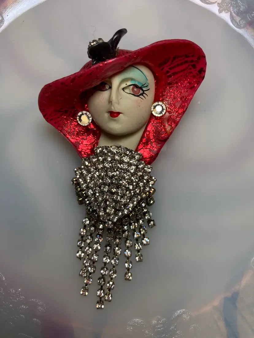 Vintage Fancy Lady Brooch Pin & Pendant. 
Hand Painted Face - LOOK AT THOSE LASHES!  Wow!
Elegant!
Large Shimmering Red Hat with a Purple Flower & Glass Stones in the Middle of It.
Décolleté is Encrusted With Faceted Prong Set Crystal Stones. 
Glass Stone Earrings.
Faceted Prong Set Crystal Stones Dangling in a Gun Metal Tone. 
Absolutely Stunning! 
Hard To Find 
Vintage 
Collectable 
Fun 
Height: 3.5” 
Width: 2”