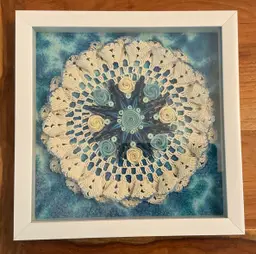 #01 Quilled blue and cream mandala on a stiffened doily. The background is batik fabric. It is in a 11 x 11” white shadowbox frame.
