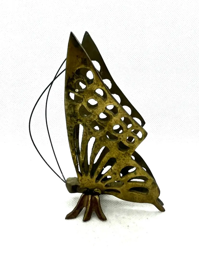This is a vintage solid brass butterfly figurine.
It is a collectible item, often dated to the 1970s.
The material is brass.
The design features a filigree butterfly silhouette with a small butterfly body at the base.
5” x 3” wide with aged patina and surface wear.