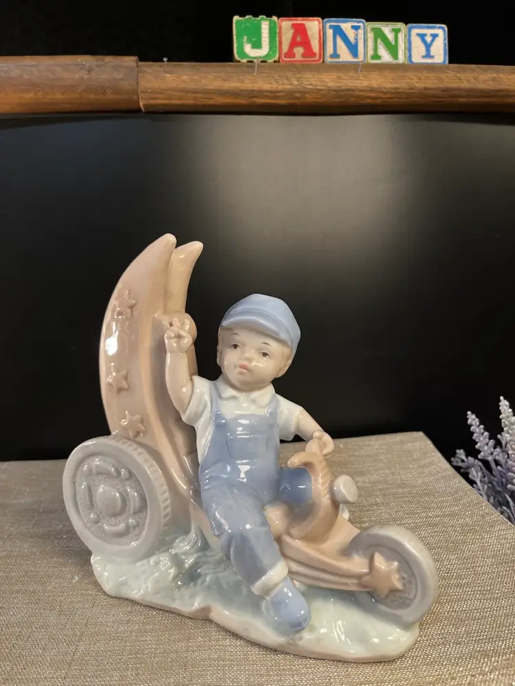 Glossy Porcelain Figurine Depicting a Young Boy in Blue Overalls & Cap, Riding a Crescent Moon that Resembles Tricycle. He is Throwing the Peace Sign!