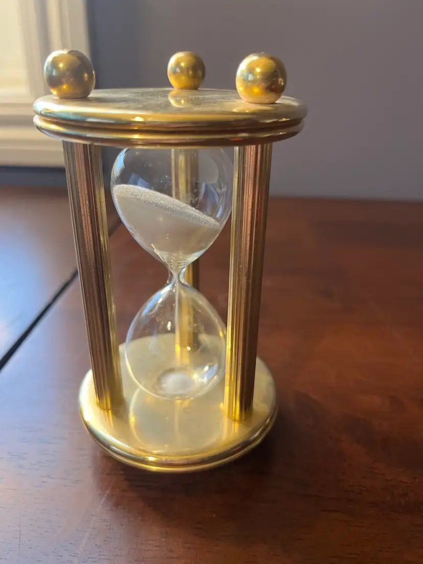 A decorative brass and glass hourglass with white sand. 5”