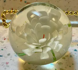 Clear glass paperweight with white flower, orange pestle, and green leaf. 2” tall and 3” wide