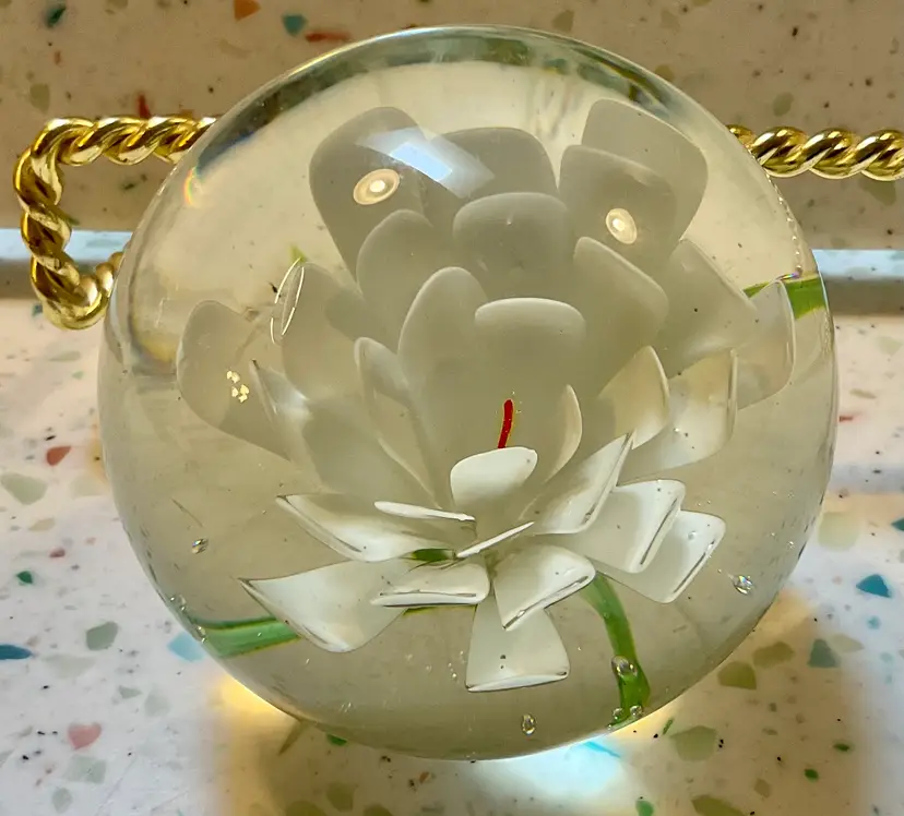 Clear glass paperweight with white flower, orange pestle, and green leaf. 2” tall and 3” wide
