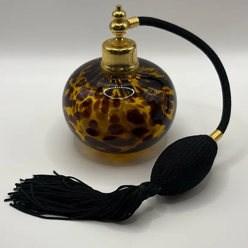 hand-blown glass perfume atomizer featuring a tortoise shell or leopard print pattern. Amber colored glass with dark brown spot patterns.
Equipped with a gold-tone metal atomizer collar and a black squeeze bulb sprayer.