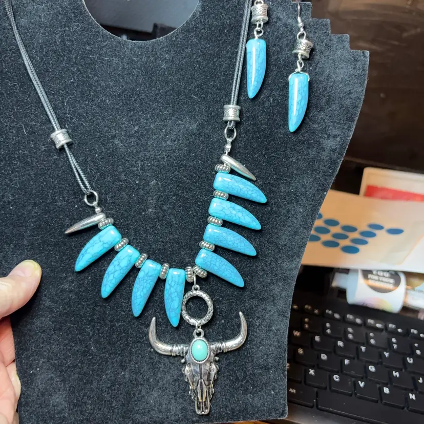 18 Inch Silvertone Necklace And Earrings Set features a central silver-tone longhorn skull pendant accented with a small turquoise stone, flanked by multiple turquoise-colored tusk or claw-shaped beads on a dark cord.