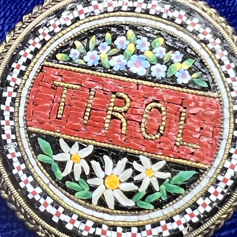 This vintage brooch is a Tiroler Mosaik item, a micro-mosaic piece from Austria featuring a brass-colored metal base and intricate craftsmanship.Marked with "Tiroler Mosaik" and an emblem depicting an eagle over stylized letters. Features a decorative rope-style border.Collectible jewelry piece. Vintage Round Micro Mosaic  With Daisey Flowers And Intricate Edge Glass 1” Wide