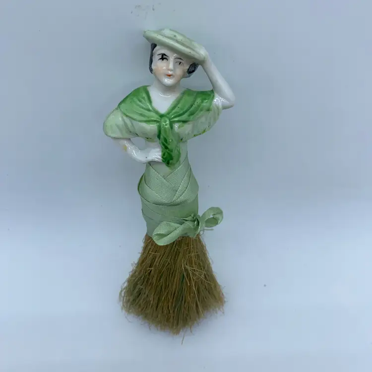 A vintage porcelain half-doll crumb brush with a straw bristle base.