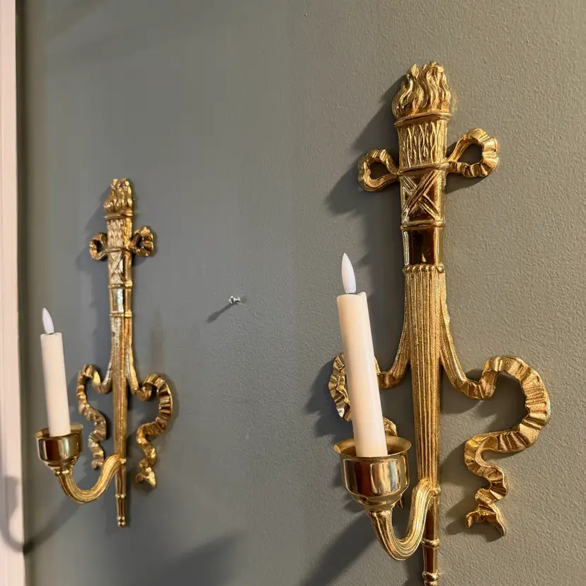 Vintage brass wall sconce Pair, often referred to as a "torch and ribbon" style candle holder. These were made by The Bombay Company, dated 1992. Elevate your home decor with these stunning vintage Bombay brass wall sconces. The intricate carvings and antique brass finish add a touch of Hollywood Regency style to any room.  (candles and votive cup not included)