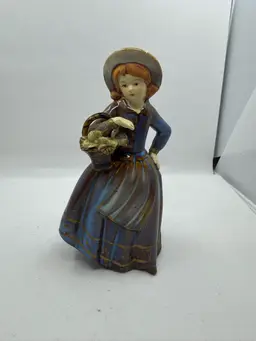 Country girl figurine holding a flower basket. Small Chip On Leaf