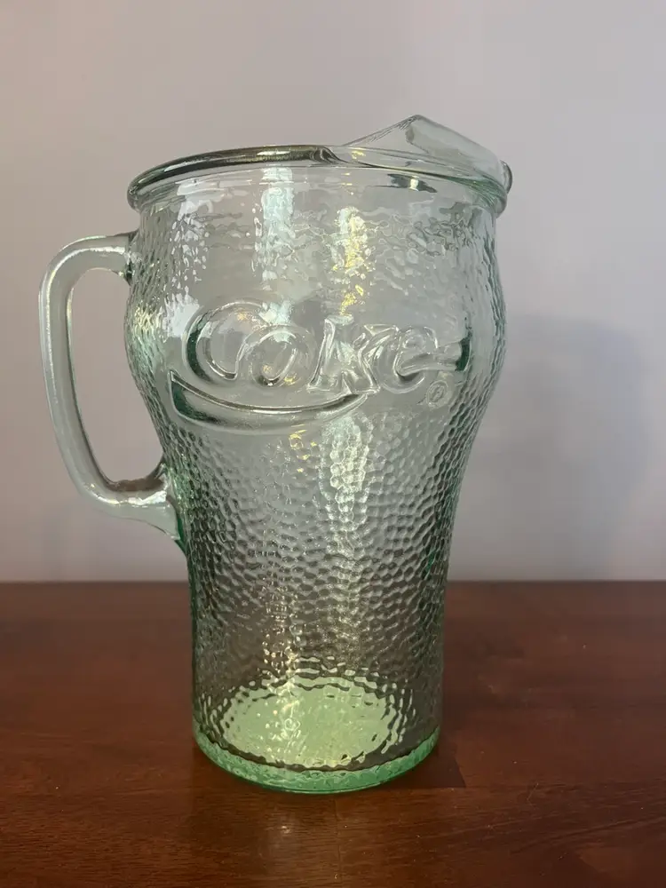A green pebbled glass Coca-Cola pitcher with an ice lip. Sticker notes, 1998. 8.5” Tall 5.5” Diameter