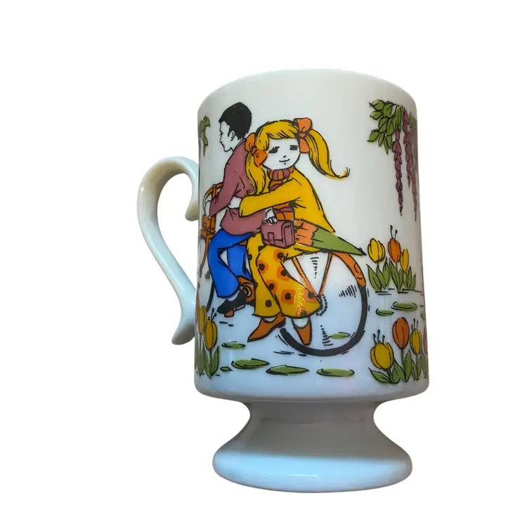 This is a vintage 1960s mod kitsch ceramic pedestal footed coffee mug with a "Boy Meets Girl" theme. 
The mug features psychedelic pop art illustrations of a girl holding an umbrella and a boy on a bicycle