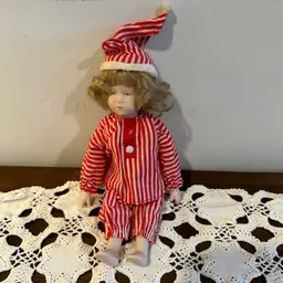 Charming vintage bisque doll dressed in red-and-white striped pajamas and a matching nightcap — a lovely collectible for doll enthusiasts and a whimsical piece of holiday decor.
This doll has a delicately painted bisque head, arms, and legs, with a soft fabric body.