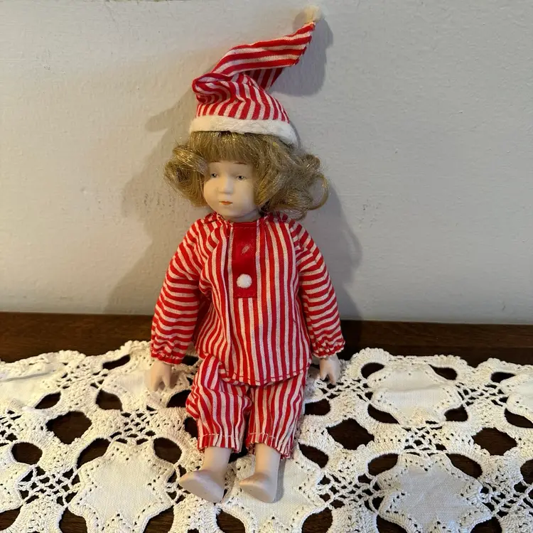 Charming vintage bisque doll dressed in red-and-white striped pajamas and a matching nightcap — a lovely collectible for doll enthusiasts and a whimsical piece of holiday decor.
This doll has a delicately painted bisque head, arms, and legs, with a soft fabric body.