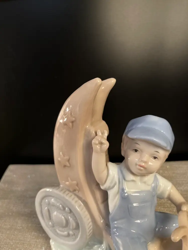 Glossy Porcelain Figurine Depicting a Young Boy in Blue Overalls & Cap, Riding a Crescent Moon that Resembles Tricycle. He is Throwing the Peace Sign!