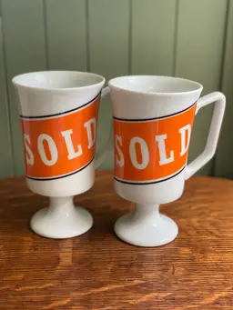 Pair Of Vintage White Pedestal Mann Made Mugs With Orange SOLD Tag On Them