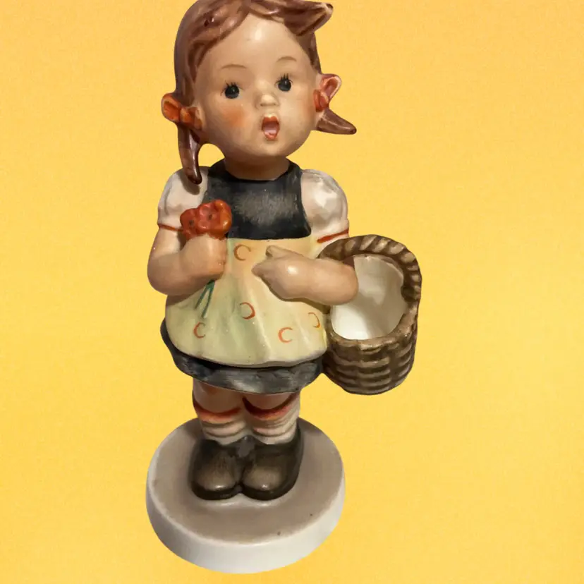 Vintage Goebel Hummel Figurine Called “ Sister” Also Know As German Name “ Schwesterlein “ Little Girl With Brown Hair & Pigtails Wearing A Yellow Apron Over A Darker Dress. Holding A Bouquet Of Orange Flowers 💐 Holding Her Little Basket 🧺. Model # 98/0 Or 98 2.0 . This Beautiful Piece Is Base On  The Artwork Of “Sister Maria Innocentia Hummel “ ( 1960)