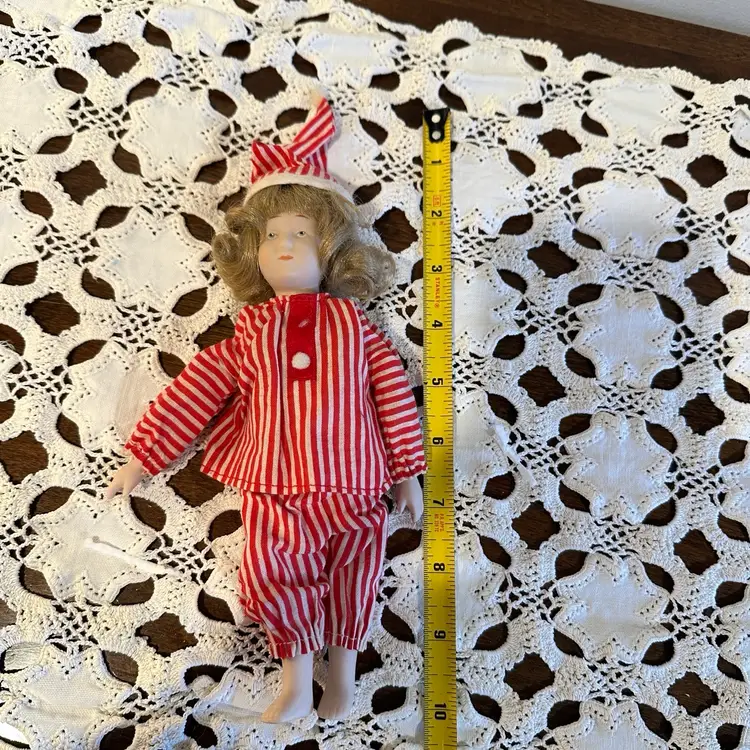 Charming vintage bisque doll dressed in red-and-white striped pajamas and a matching nightcap — a lovely collectible for doll enthusiasts and a whimsical piece of holiday decor.
This doll has a delicately painted bisque head, arms, and legs, with a soft fabric body.