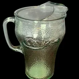 A green pebbled glass Coca-Cola pitcher with an ice lip. Sticker notes, 1998. 8.5” Tall 5.5” Diameter