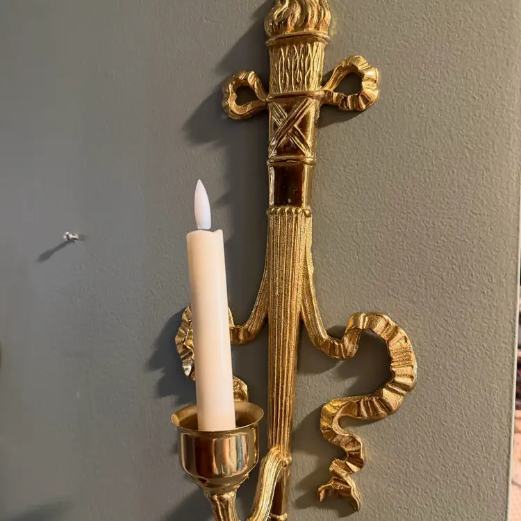 #01 Vintage brass wall sconce Pair, often referred to as a "torch and ribbon" style candle holder. These were made by The Bombay Company, dated 1992. Elevate your home decor with these stunning vintage Bombay brass wall sconces. The intricate carvings and antique brass finish add a touch of Hollywood Regency style to any room.  (candles and votive cup not included)