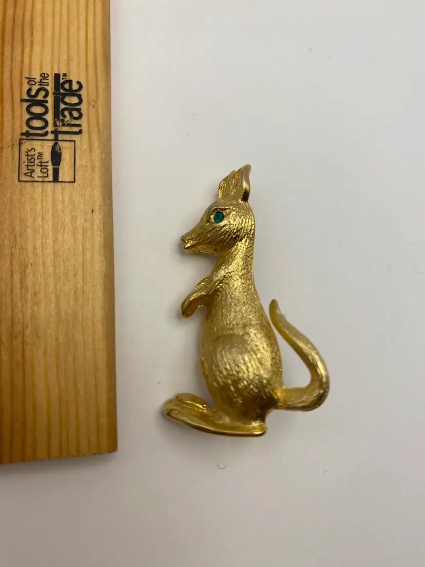 It's a gold-toned metal pin in the shape of a kangaroo, featuring a textured surface and a green rhinestone eye. 1990’s