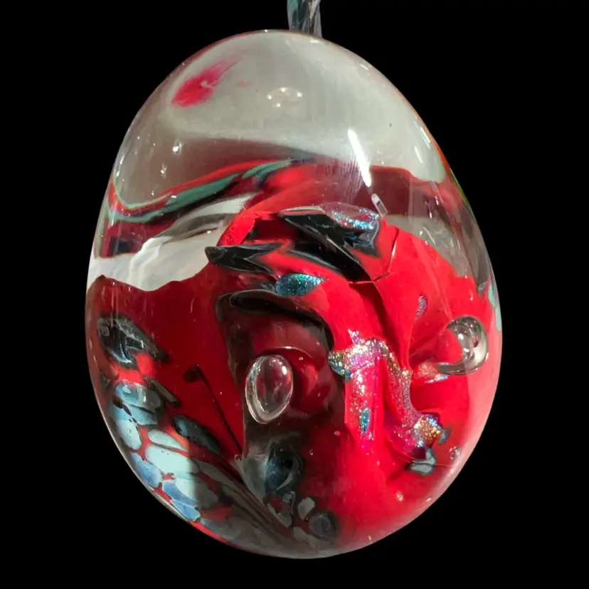 an egg-shaped art glass paperweight designed by the renowned American glassblower Rollin Karg. Rollin Karg is a prominent Kansas-based artist celebrated for his masterfully crafted studio art glass. His work is easily distinguished by thick clear glass encasing highly vibrant, fluid interiors often accented with metallic finishes, floating controlled bubbles, and shimmering dichroic glass flakes. Pieces from this specific line feature a signature hand-etched on the base or lower edge