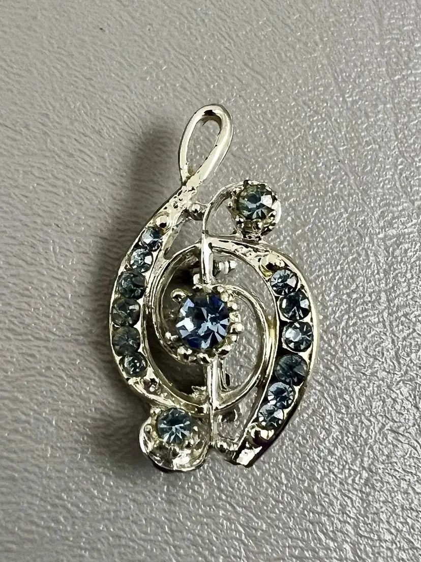 This brooch is for anyone whose whole personality is music, and their coat should say so too.  It’s a silver-tone treble clef, dressed up with a row of light blue rhinestones down each curve, a bigger stone in the center, and one on each end. It’s the kind of pin that goes on a choir robe, a piano teacher’s cardigan, or a band mom’s jacket at the winter concert.  All the stones are present and still catch the light nicely. A few are a little cloudy, the way vintage glass gets after years of applause. The back has some small tarnish spots that nobody will ever see unless they’re sitting really close during the slow movement. The pin closes securely.  It measures about 3/4" wide and 1-5/8" tall, big enough to notice and small enough to stay classy.  It’s a great gift for musicians, music teachers, choir directors, or the kid who finally made first chair. Please check the photos, since they’re part of the description.