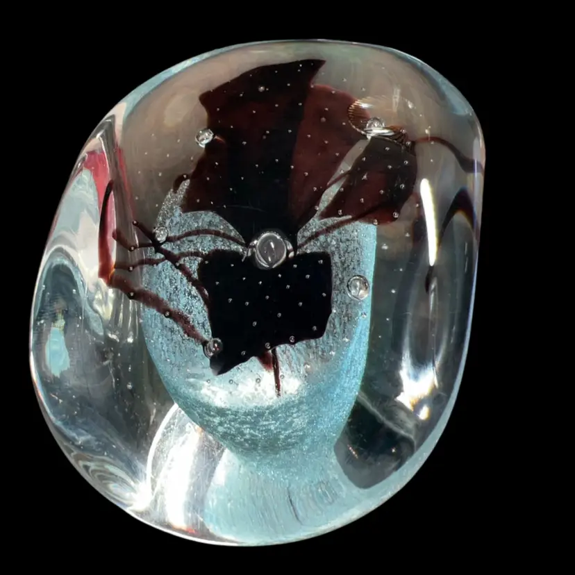 • Artist: Michael Nourot, an influential figure in the American Studio Glass movement who co-founded Nourot Glass Studio in Benicia, California, in 1974.
• Design Motifs: This paperweight utilizes his signature clear crystal encasement housing a deep, dark burgundy/oxblood abstract flower structure suspended over a shimmering controlled-bubble background.
• Markings: The code inscribed next to the signature (such as CN 50-88) serves as the studio's internal tracking registry, indicating the specific series design and the year it was hand-blown (1988).